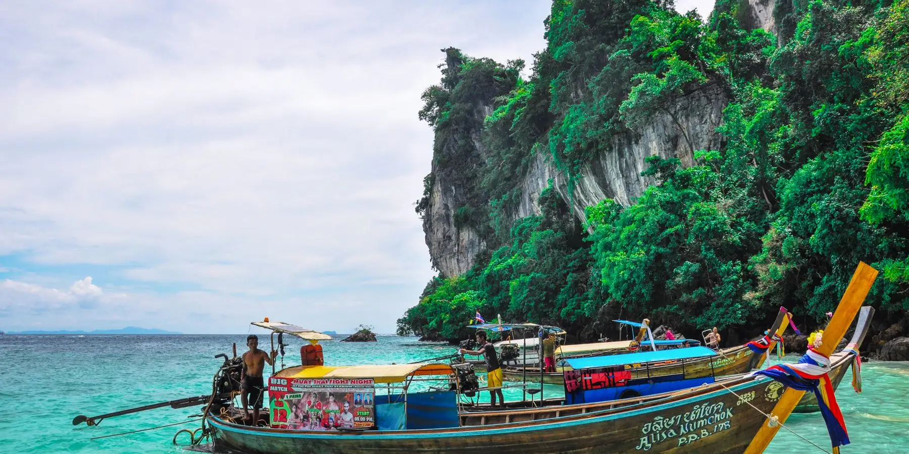 Traditional Thai longtail boats moored in turquoise shallows beneath a forested limestone cliff.