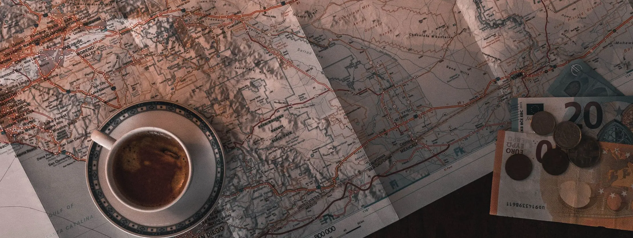 A folded paper map on a desk with a cup of coffee, loose coins and banknotes resting on one corner.